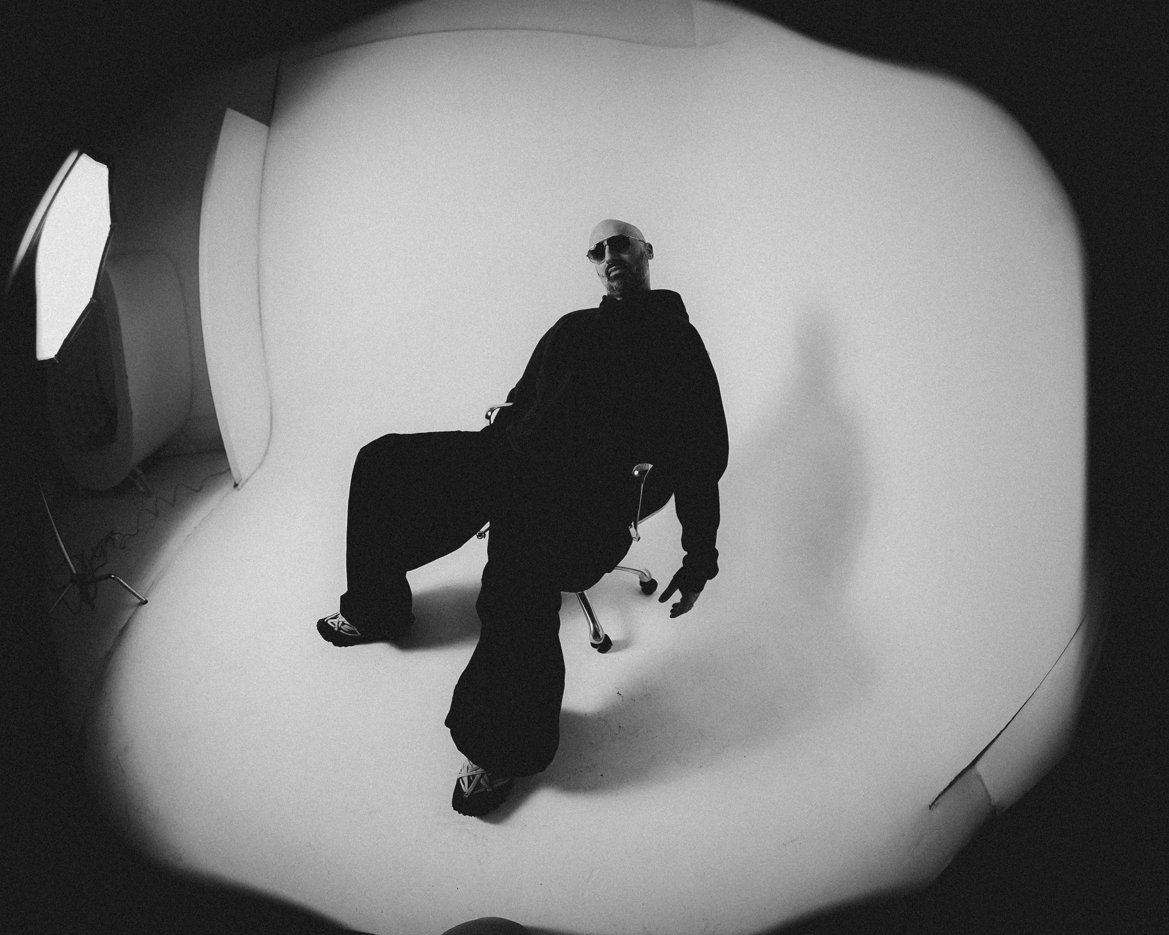 DECAP seated in an office chair on a white cyc, all black, sunglasses, shot wide on a fisheye in black and white. Photograph by Shaina Rose.